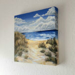 Acrylbild, das Sanddünen mit Gras vor einen blauen Meer zeigt, darüber ein blauer Himmel mit weißen Wolken, vor einer weißen Wand in der Seitenansicht gezeigt